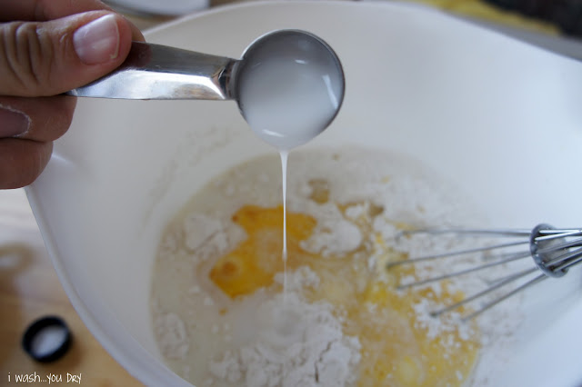 A hand holding a measuring spoon over a bowl of needed ingredients for vanilla donuts