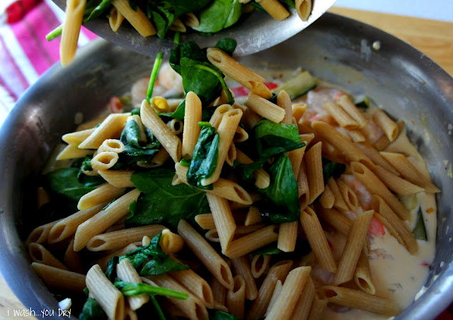 Penne pasta and spinach leaves being added to a pan of cream sauce and veggies.