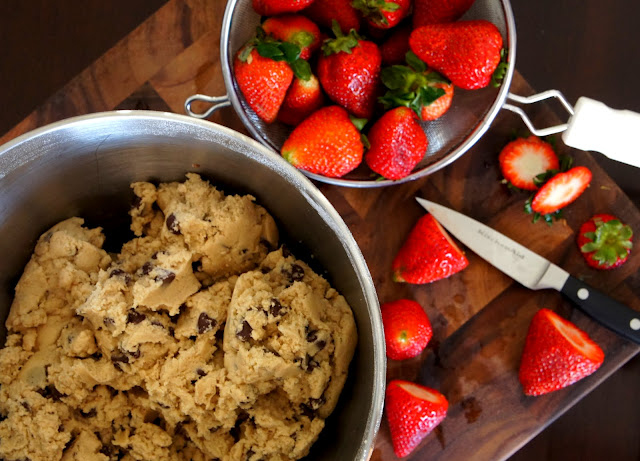 A cutting board with a knife cutting strawberry tops off, next to with a bowl of raw cookie dough, and strainer strawberries 