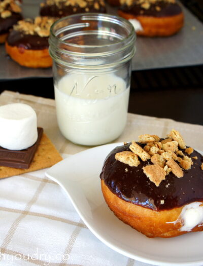 A Marshmallow Cream filled S'mores Doughnut displayed on a plate next to a glass of milk and an uncooked S'more