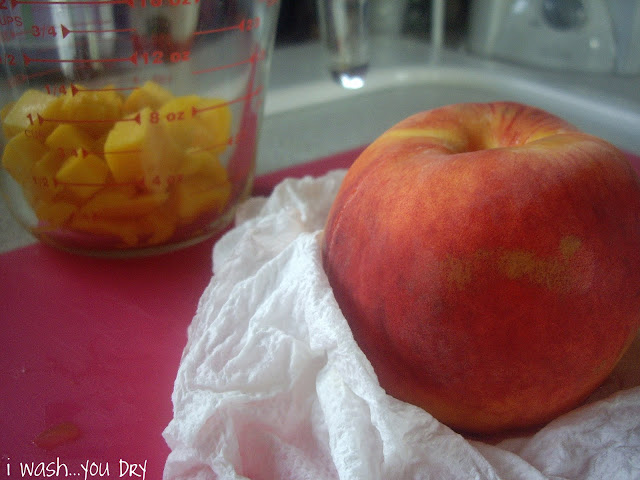 A peach on a cutting board with a glass measuring cup with cubbed mango pieces in it. 