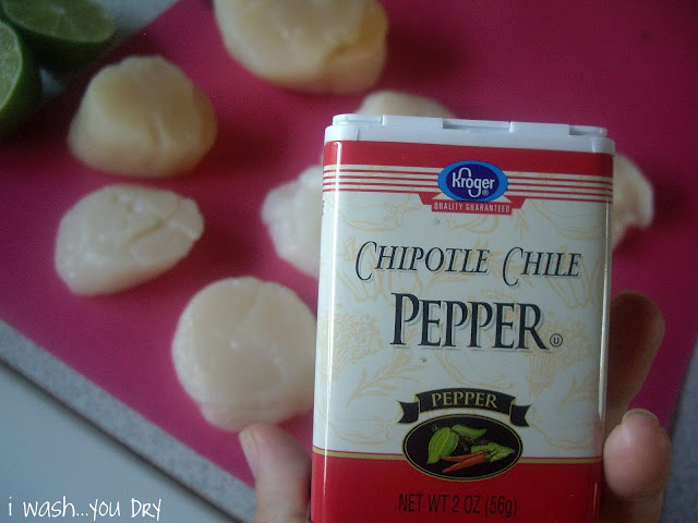 A hand holding a container of chipotle Chile pepper above sliced scallops on a cutting board. 