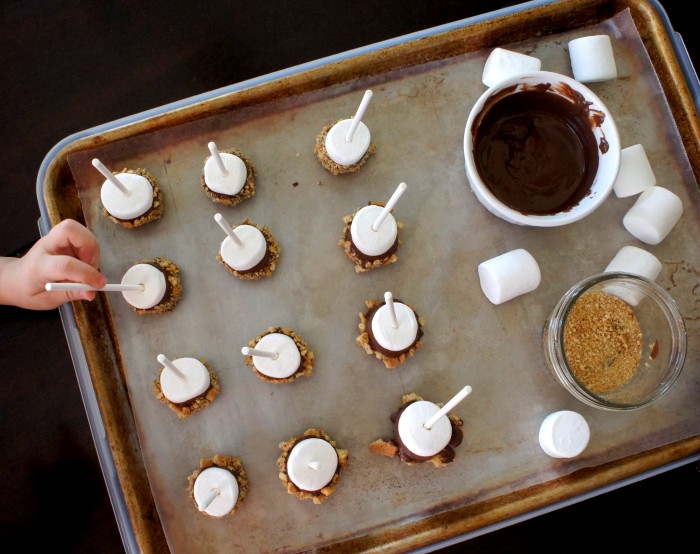 A look down on a pan of marshmallow pops drying on a pan next to a bowl of chocolate sauce and graham cracker crumbs