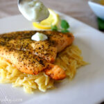 A spoon dripping Tzatziki Sauce on a Greek Salmon Fillet on a bed of noodles.