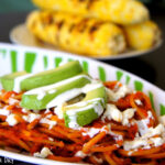 A bowl of red pasta with slices of avocado on top drizzled with a white sauce