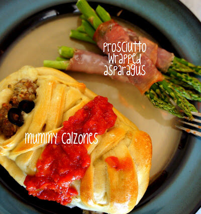 A mummy shaped calzone, topped with a little sauce on a plate next to bacon wrapped asparagus