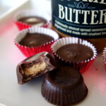 Chocolates on a table in front of a jar of cookie butter, one of the chocolate has a bite taken from it