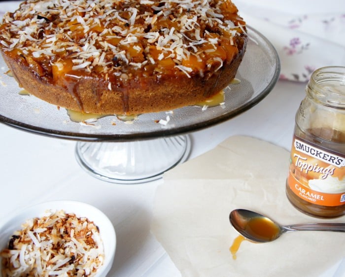 An upside down mango cake displayed on a clear glass cake plate  topped with caramel and coconut flakes