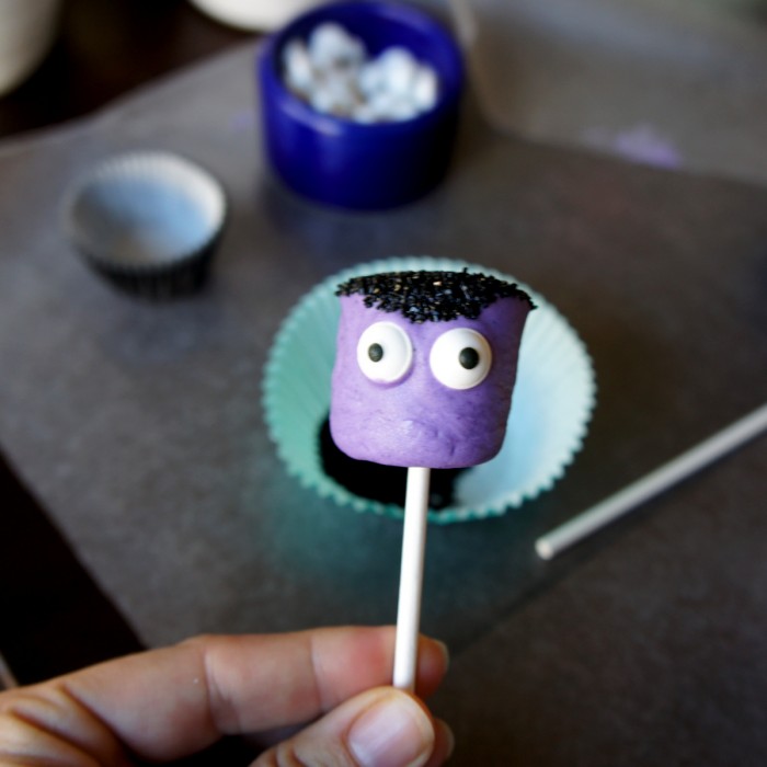 The front view of a purple dipped marshmallow pop with black sprinkles for hair and two candied eyes added