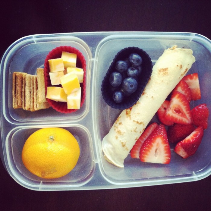 A plastic container filled with food, with easy bento lunch ideas - Cheese, crackers, fruit and a rolled tortilla