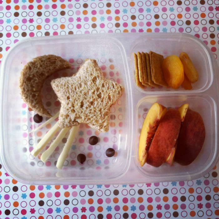 A Bento styled plastic container with dividers showcasing an easy lunch of a star shaped sandwich shaped to look like a shooting star, crackers and peach slice