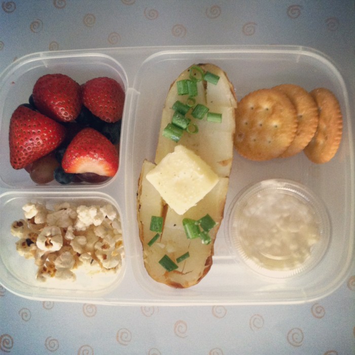 A bento box filled with strawberries, popcorn, half a baked potato with butter on top, chips and dip