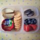 A Bento styled plastic container with dividers showcasing an easy lunch of crackers, peanut butter, apples, cookies and fruit