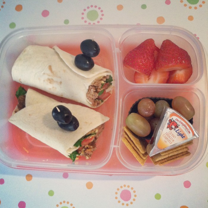 A bento box filled with two halves of a wrap, berries, grapes, crackers and a cheese wedge in the compartments