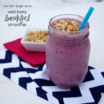 A smoothie in a glass cup with granola on top and a blue straw next to a small bowl of granola