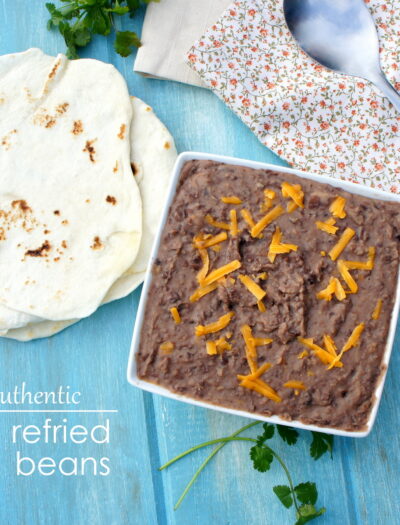 A square bowl of refried beans topped with cheese next to a small pile of tortillas