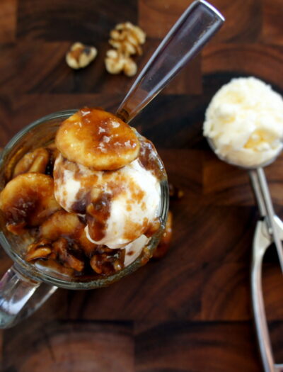 A look on from above of a glass mug filled vanilla ice cream topped with Banana Fosters and a ice cream scooper with vanilla ice-cream next to it