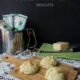 Three biscuits on a wooden cutting board surrounded by tubes of herbs and a block of cheese