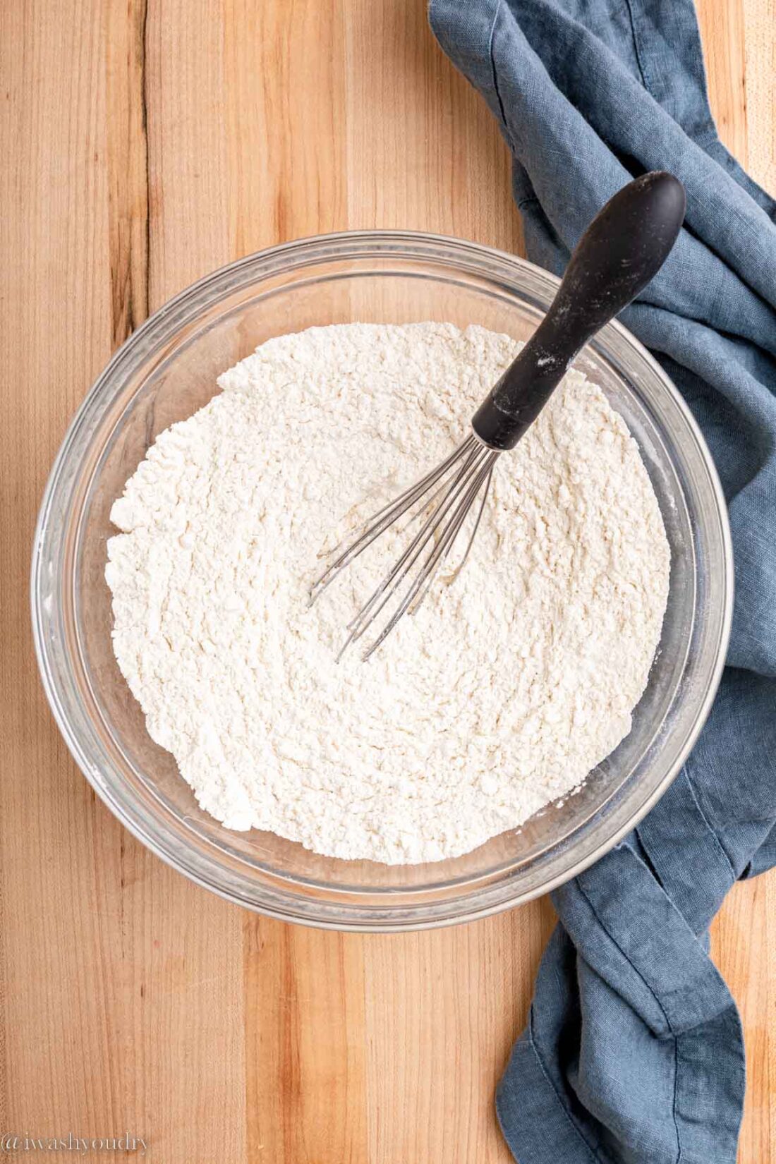 Flour mixture in glass bowl with wire whisk near blue dish towel. 