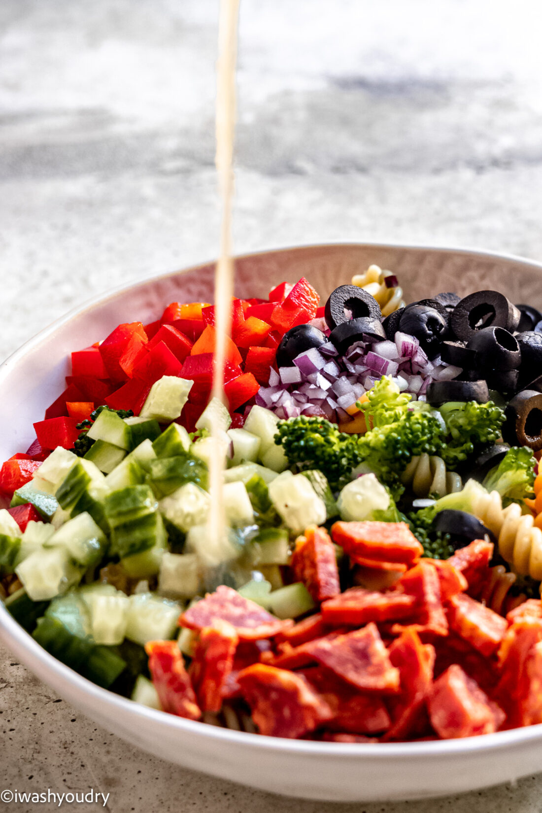 Pouring italian dressing into pasta salad with pepperoni and broccoli.