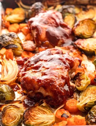 Baked bbq chicken on metal sheet pan with carrots and brussels sprouts.