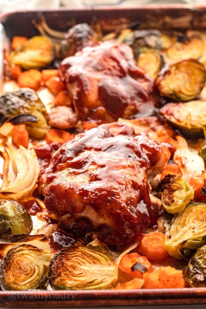 Baked bbq chicken on metal sheet pan with carrots and brussels sprouts.