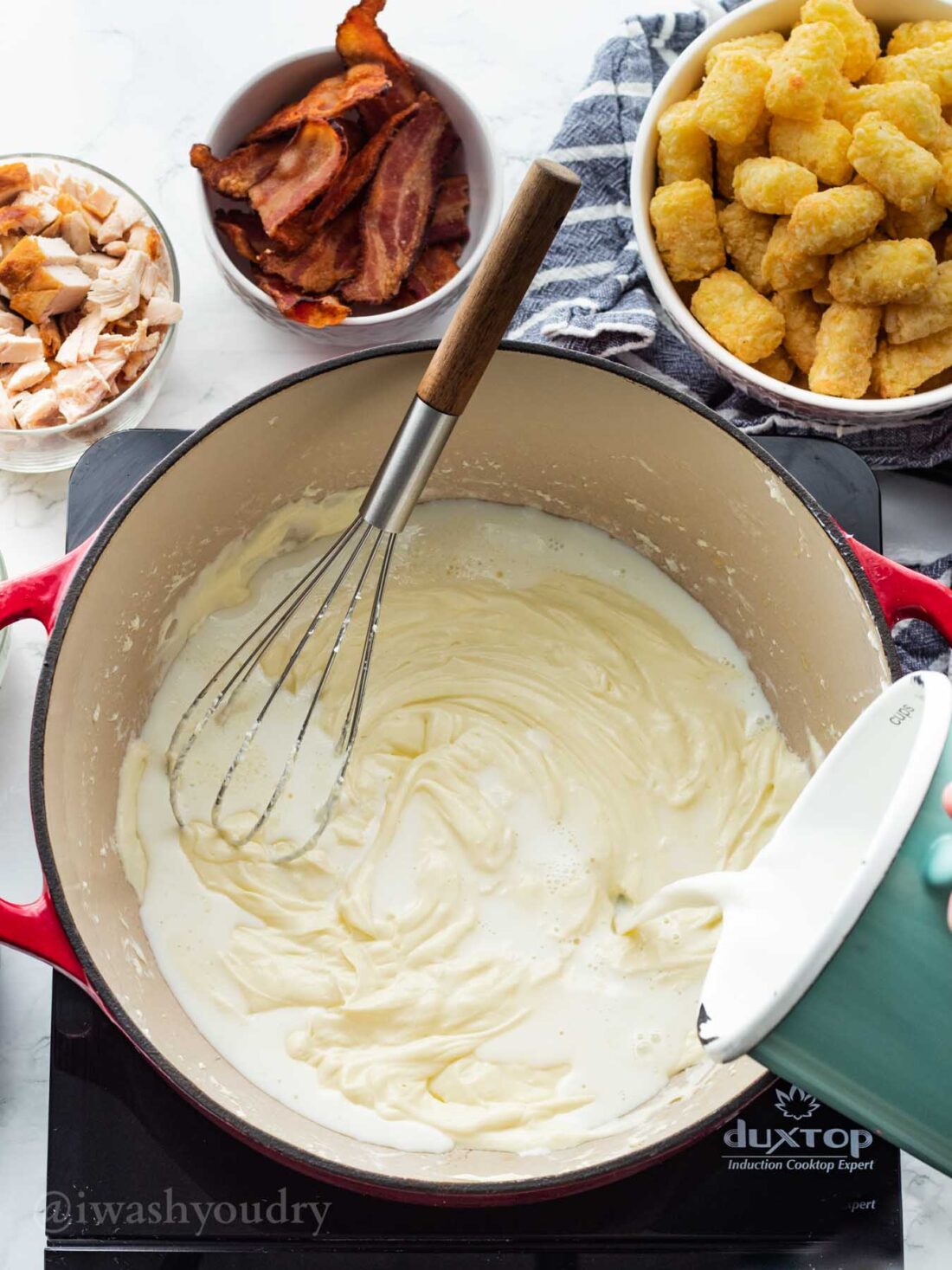 Pouring blue pitcher of milk into pot of melted butter and cream cheese with whisk. 