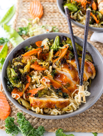 A bowl of food, with chicken, veggies and ramen noodles and a pair of chopsticks