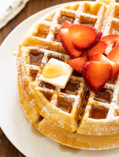 Crispy waffles on white plate with syrup and strawberries.