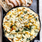 Black pan of spinach artichoke dip with bread on side.