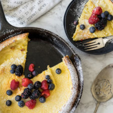 Skillet of food with a plate next to it with a slice of pancake topped with raspberries and blueberries