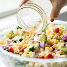 A container of food on table, with Pasta and veggies being topped with a dressing from a jar