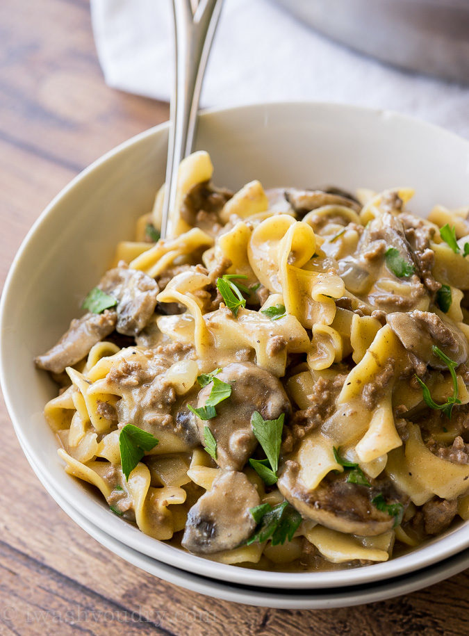 A close up of a bowl of food on a plate, with beef, noodles and a white sauce