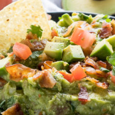 A close up of guacamole with a chip, bacon and tomatoes