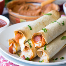 A plate full of food, with stacked taquitos on it and hummus in the background