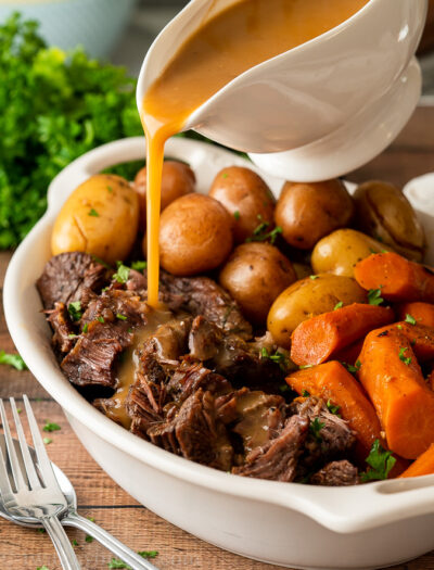 Pouring gravy over shredded beef pot roast in white dish.