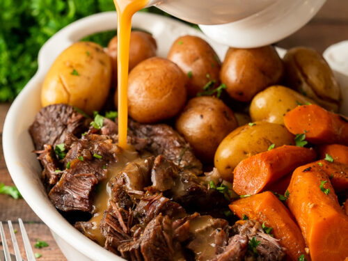 Pouring gravy over shredded beef pot roast in white dish.