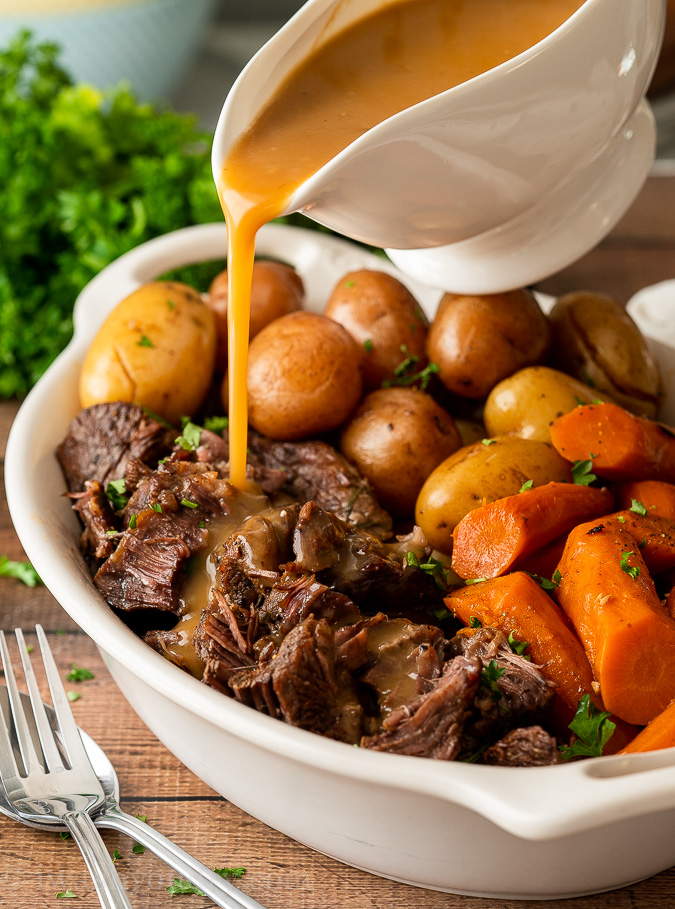 Pouring gravy over shredded beef pot roast in white dish.