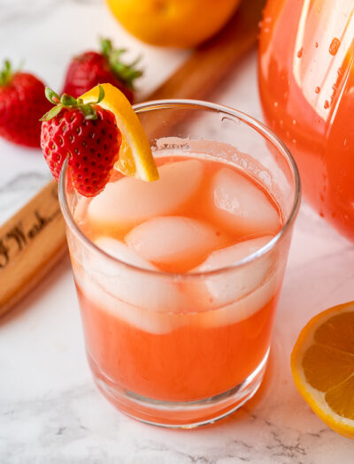 Clear glass full of strawberry lemonade with ice. Garnished with fresh strawberries and a lemon slice.