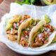 Three fish tacos on a plate, topped with avocado, diced tomato, onion and cilantro.