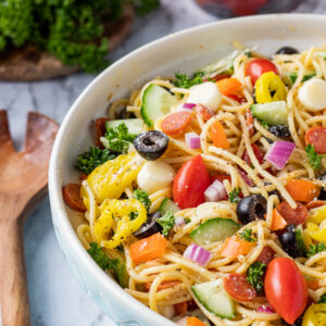 Blue bowl with spaghetti, tomatoes, cucumbers, peppers, olives, and italian dressing on marble table with wood spoon.