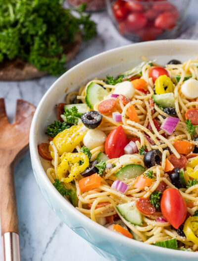 Blue bowl with spaghetti, tomatoes, cucumbers, peppers, olives, and italian dressing on marble table with wood spoon.