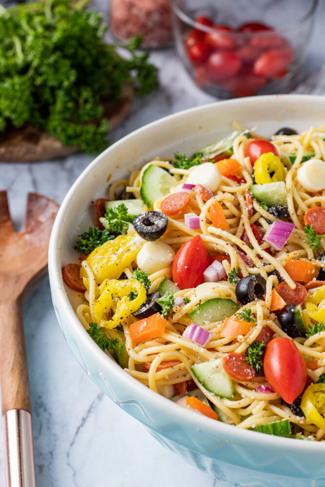 Blue bowl with spaghetti, tomatoes, cucumbers, peppers, olives, and italian dressing on marble table with wood spoon.