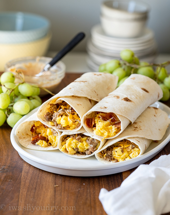 Breakfast burritos on a plate with grapes and sauce on side.