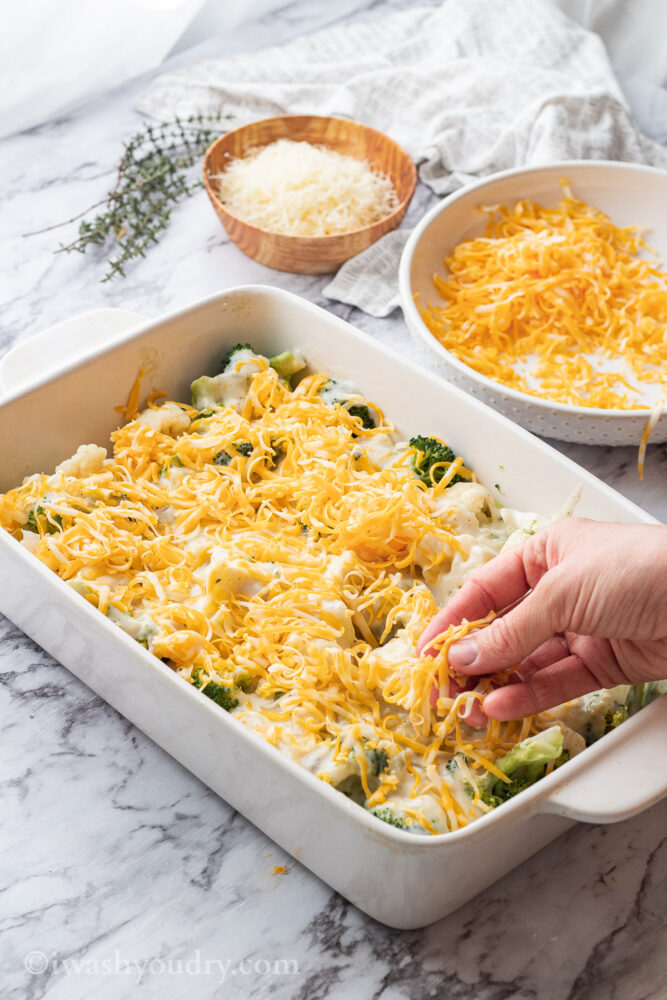 Sprinkling cheese over white pan of broccoli cauliflower casserole. 