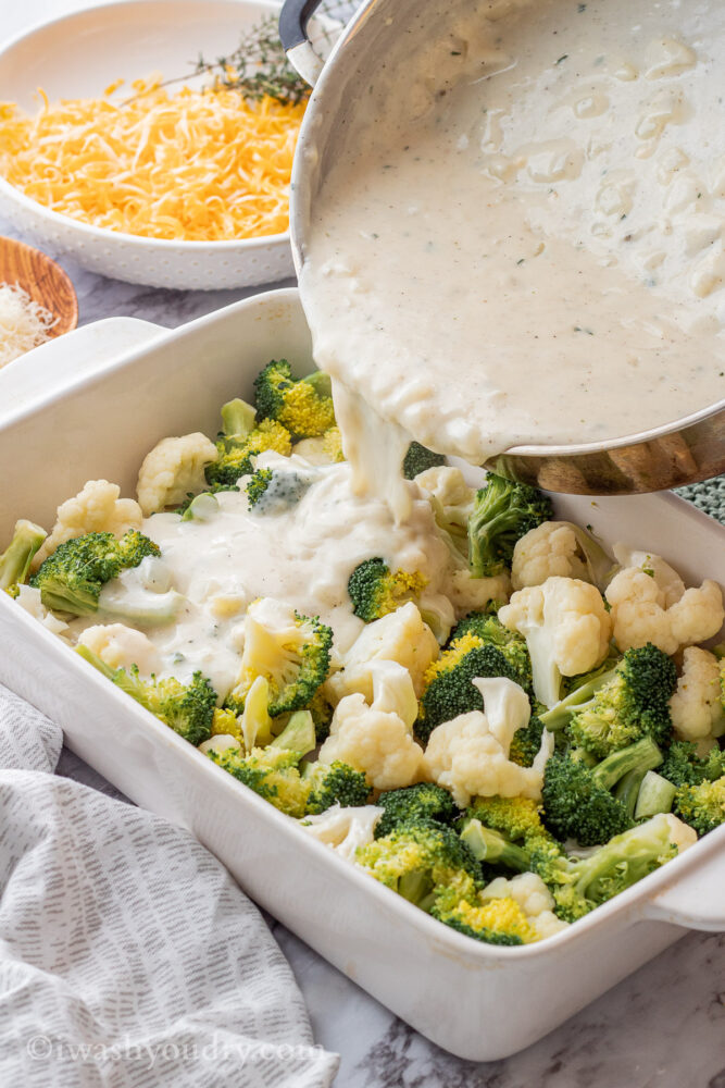 Pouring creamy sauce into white pan of broccoli of cauliflower. 