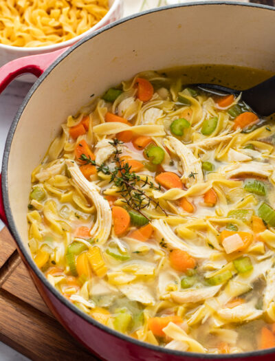 Cooked Chicken Noodle Soup in red pot on wood cutting board.