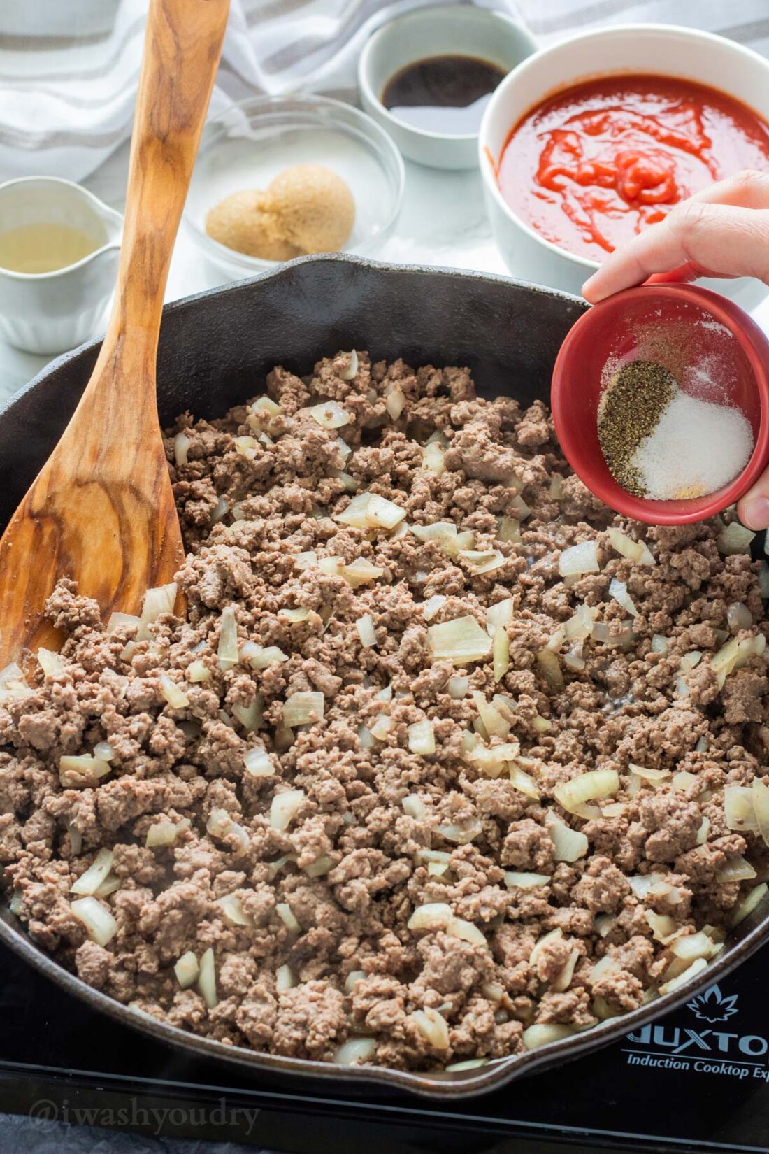 Pouring red bowl of spices into a black frying pan of cooked ground beef. 