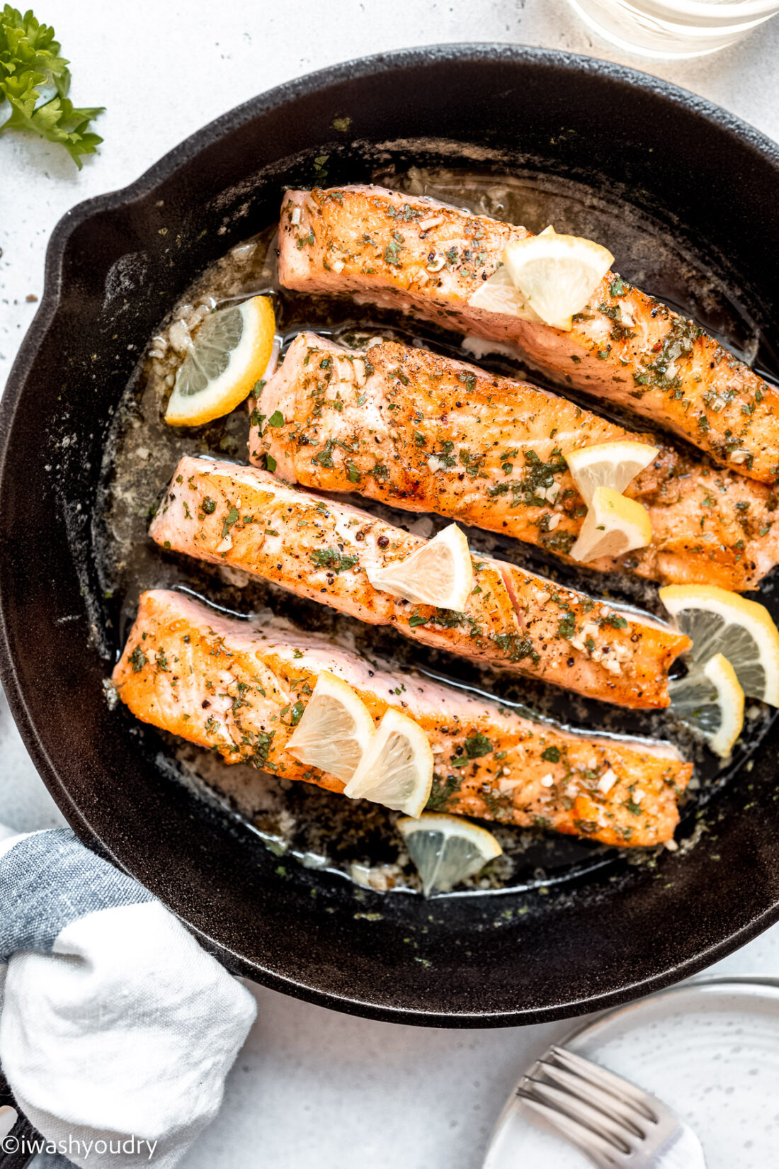 Cooked lemon garlic salmon in black pan with parsley and lemon slices with plates and fork to the side. 