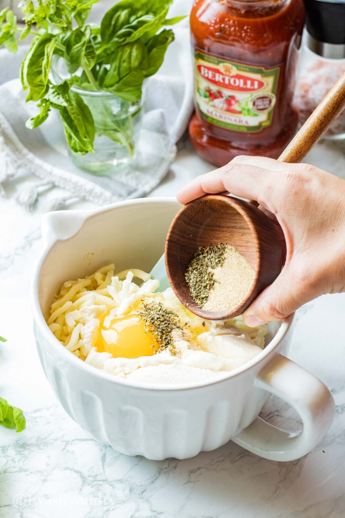 Wood bowl of spices pouring into white bowl of ricotta, mozzarella, parmesan, and egg. 